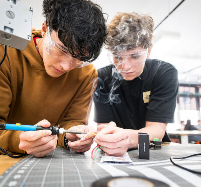 Two students wear eye protection and rest their arms on a work table as they solder connections on a sensor.