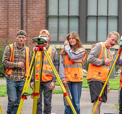 Eight students stand outside campus buildings in front of surveying equipment with a faculty member providing instruction.