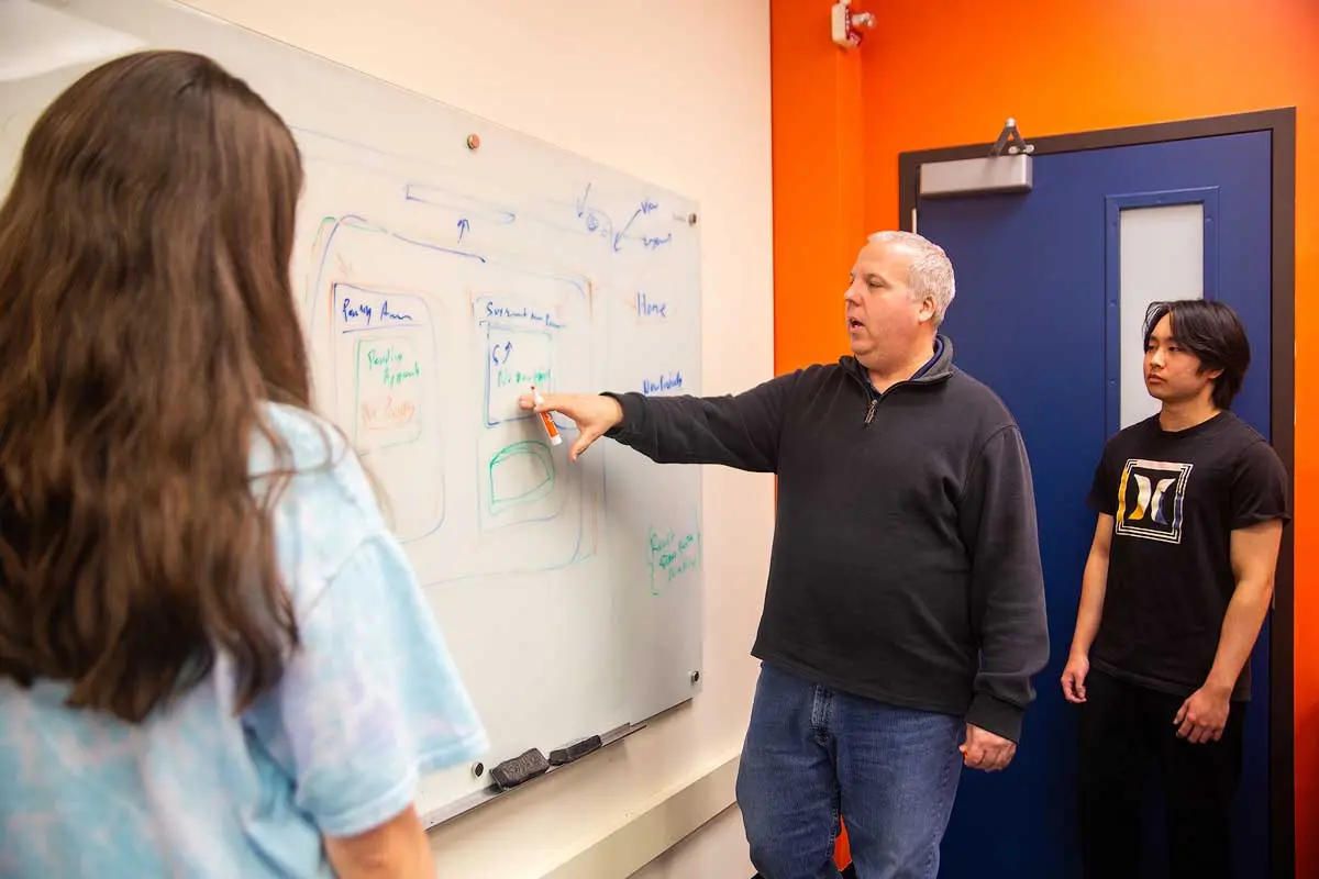 A person writes on a whiteboard standing in front of two students.